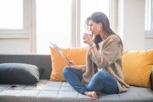 Young woman sitting on the sofa drinking coffee and working on the digital tablet